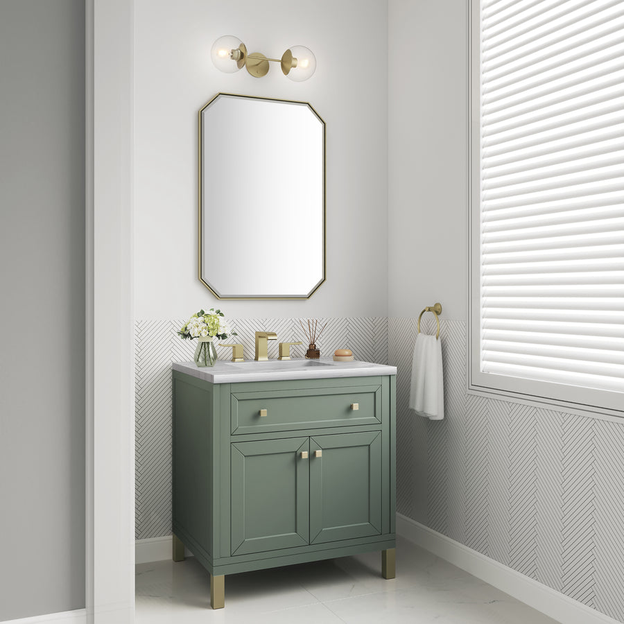 A small bathroom corner with a green vanity, white countertop, flowers in a vase, a gold-framed mirror, gold fixtures, a white towel on a gold ring, and a window with closed white blinds.