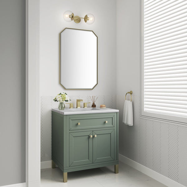 A small bathroom corner with a green vanity, white countertop, flowers in a vase, a gold-framed mirror, gold fixtures, a white towel on a gold ring, and a window with closed white blinds.