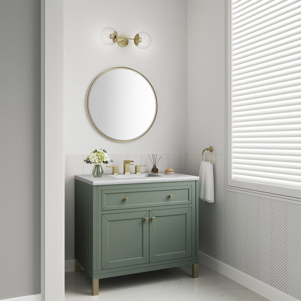 A modern bathroom vanity with a green cabinet, white countertop, round mirror, and brass fixtures. A vase with flowers and toiletries sit on the counter; a towel hangs by a window with closed blinds.