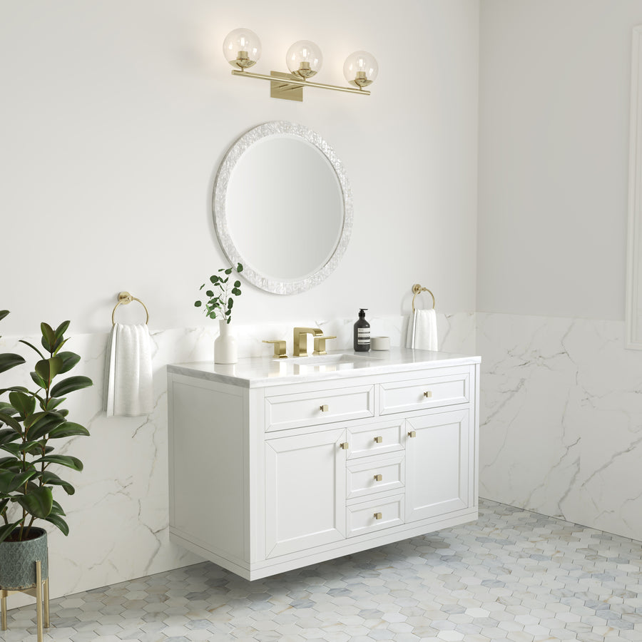A modern bathroom with a white floating vanity, round mirror, gold fixtures, marble walls, hexagon floor tiles, a gold light fixture above the mirror, and green potted plants for decoration.