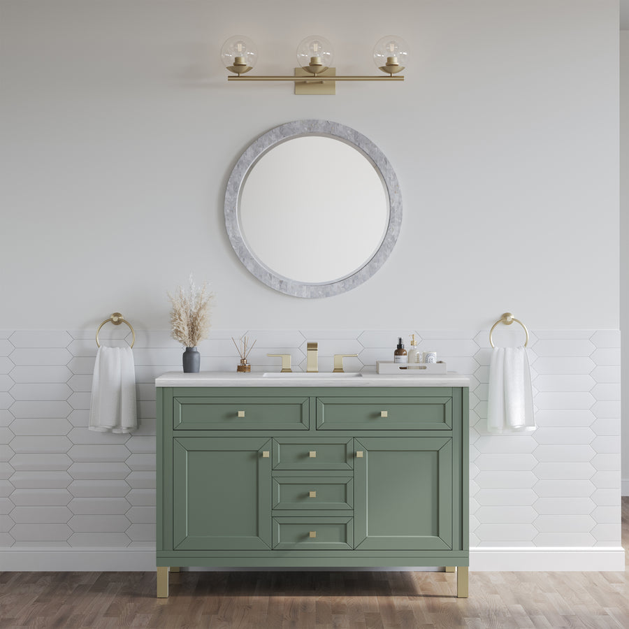 A modern bathroom includes the Chicago 48" Vanity in sage green with gold hardware, a round mirror, a gold light fixture, hexagonal white tile backsplash, two hand towels on gold rings, and wood flooring.