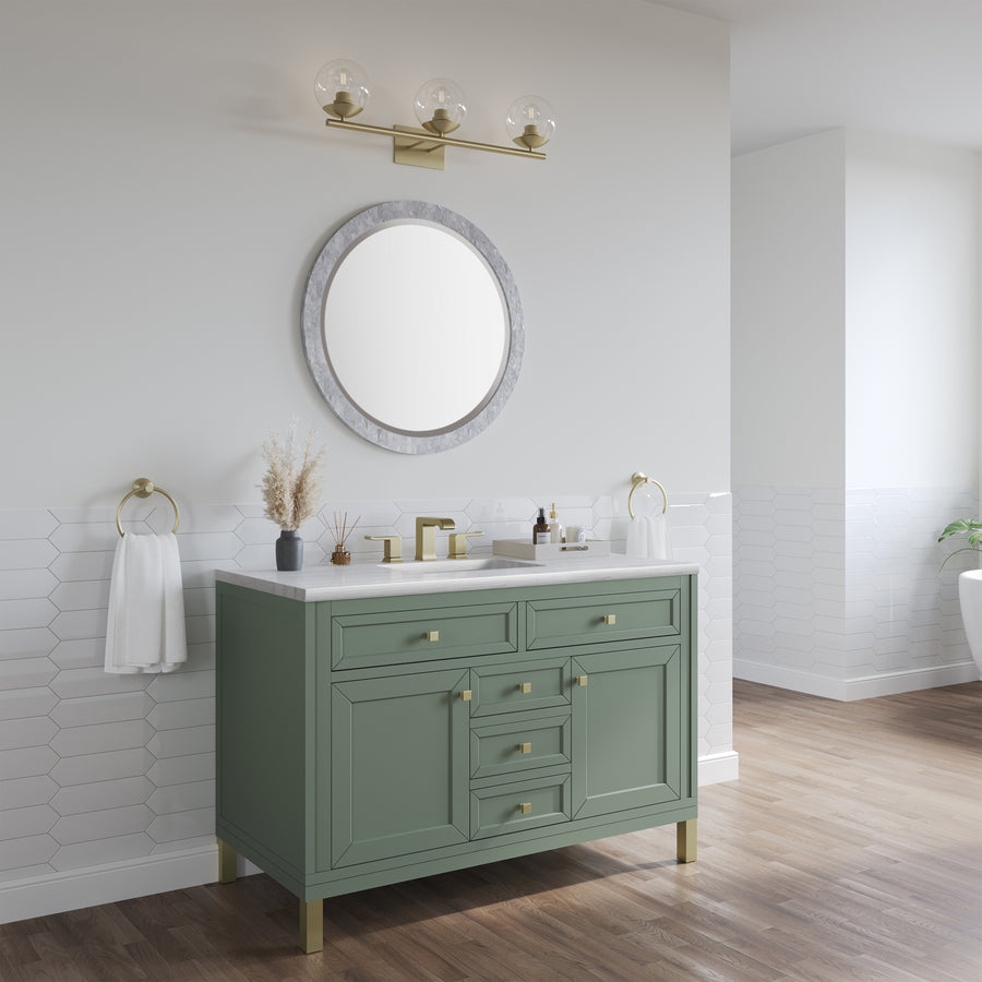A modern bathroom features a green vanity with gold hardware, a round mirror, gold light fixture, white countertop, towel rings, and white hexagonal wall tiles above wood flooring.