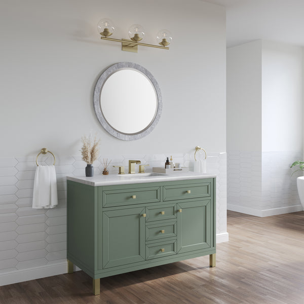 A modern bathroom features a green vanity with gold hardware, a round mirror, gold light fixture, white countertop, towel rings, and white hexagonal wall tiles above wood flooring.
