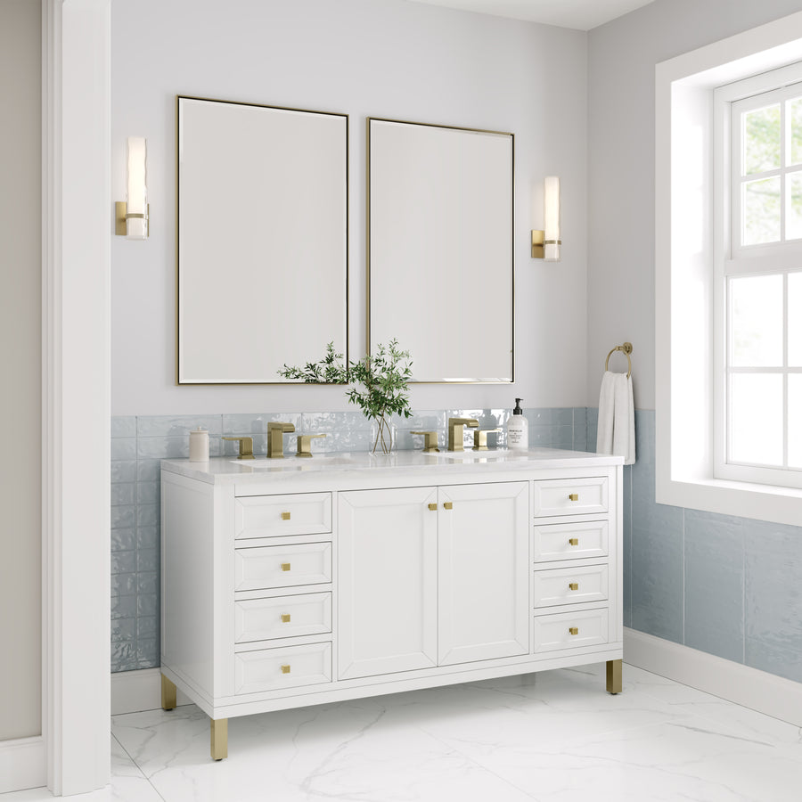 Modern bathroom with a white vanity, gold accents, dual mirrors, wall sconces, marble floor, blue half-wall tiles, and a window letting in natural light; small plant and toiletries sit on the countertop.