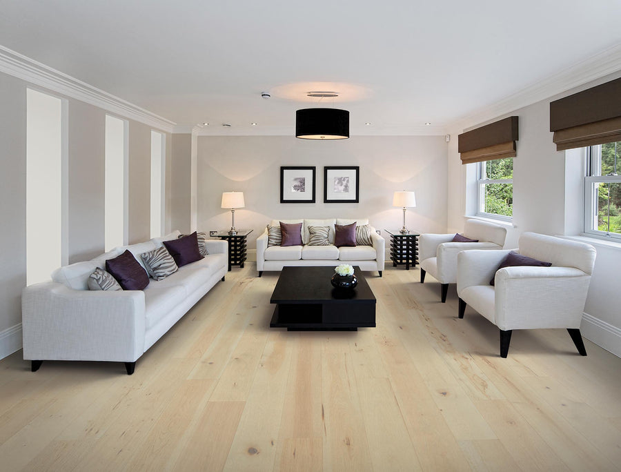 A modern living room with light wood flooring, white sofas and armchairs, a black coffee table, two table lamps, framed art on the wall, and large windows with brown blinds.