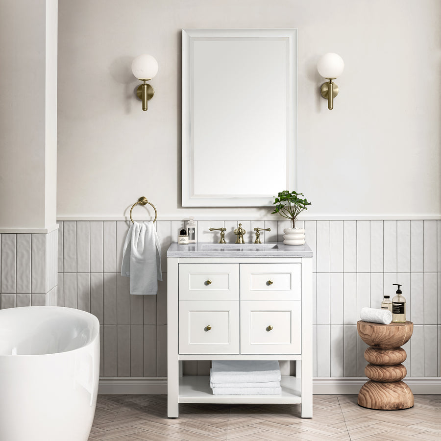 A transitional bathroom featuring the Breckenridge 30" Vanity with a marble countertop, square mirror, wall sconces, potted plant, white towel, toiletries, decorative wooden stool, and a partial view of a white bathtub.
