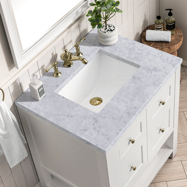 A white bathroom vanity with a marble countertop, gold faucet, potted plant, soap dispenser, and decorative items, next to a wooden stool holding a soap dish and rolled towel.