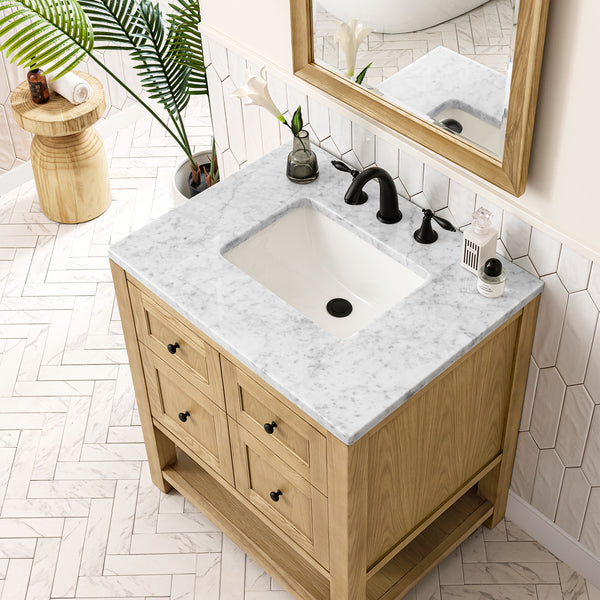 A modern bathroom vanity with a white marble countertop, black faucet, wooden cabinet, decorative items, and a mirror above, set on a light herringbone tile floor with a plant nearby.