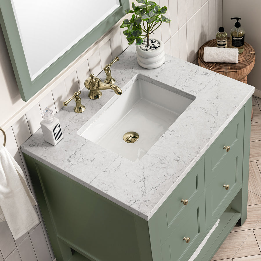 A bathroom vanity with a white and gray marble countertop, sage green cabinet, gold faucet, potted plant, soap dispenser, and toiletries on a small wooden stool beside it.
