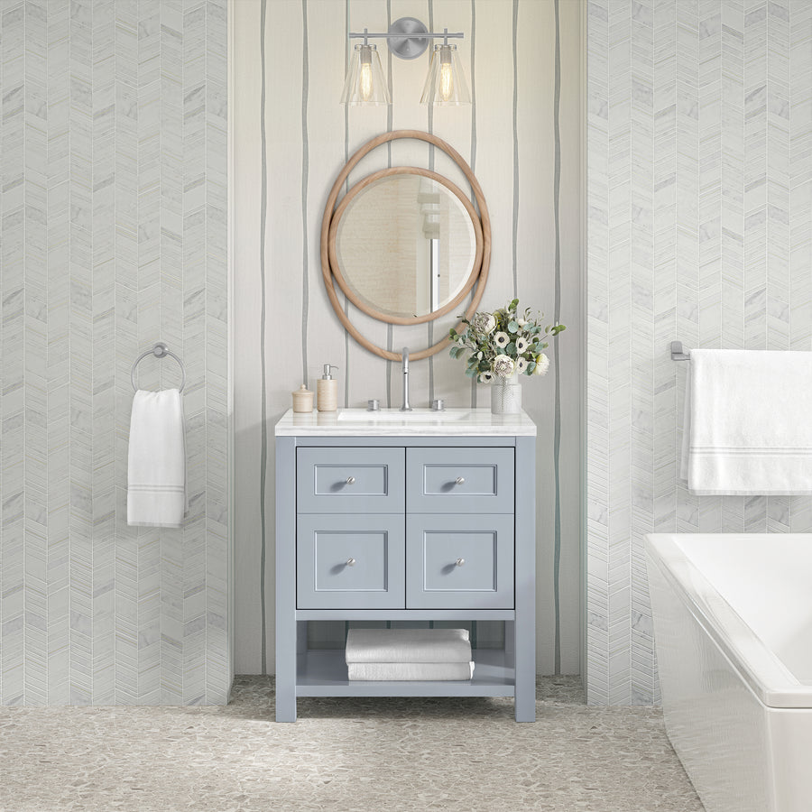 A modern bathroom features the Breckenridge 30" Vanity in light blue, a round mirror, double sconces, white countertop, and flower vase. White towels hang on both walls; a bathtub sits right. Pale patterned wallpaper enhances the transitional style.