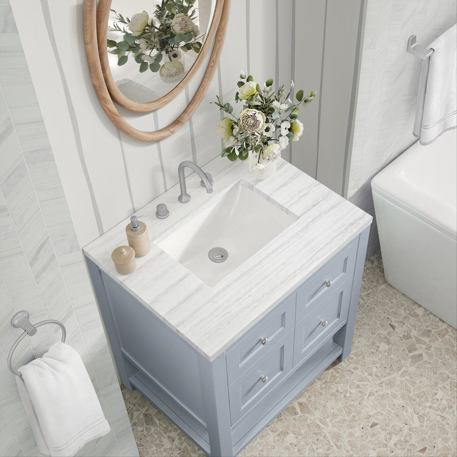 Overhead view of a bathroom vanity with a white marble countertop, silver faucet, light blue drawers, a round mirror, a bouquet of flowers, soap dispensers, and a towel hanging on the wall next to a bathtub.