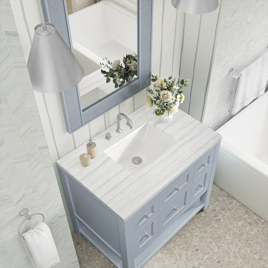 Overhead view of a bathroom vanity with a white marble countertop, light blue cabinet, sink, faucet, soap dispenser, vase with white and green flowers, mirror, and part of a bathtub visible.
