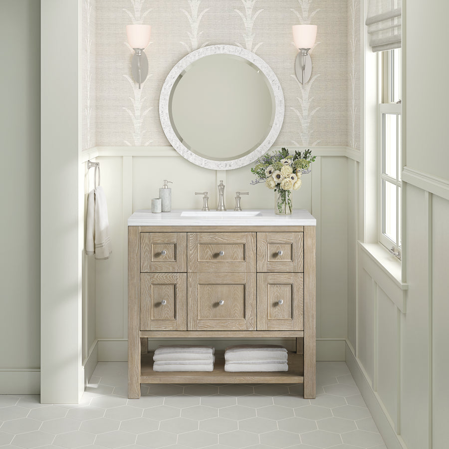 The Breckenridge 36" Vanity with a white countertop, round mirror, wall sconces, flowers, soap dispensers, and towels below adds transitional style to this bright bathroom.