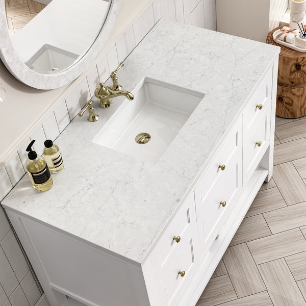 White marble bathroom vanity with an under-mount rectangular sink, brass faucet, and matching brass hardware. Soap dispensers sit on the counter, with a round mirror and light wood herringbone floor visible.