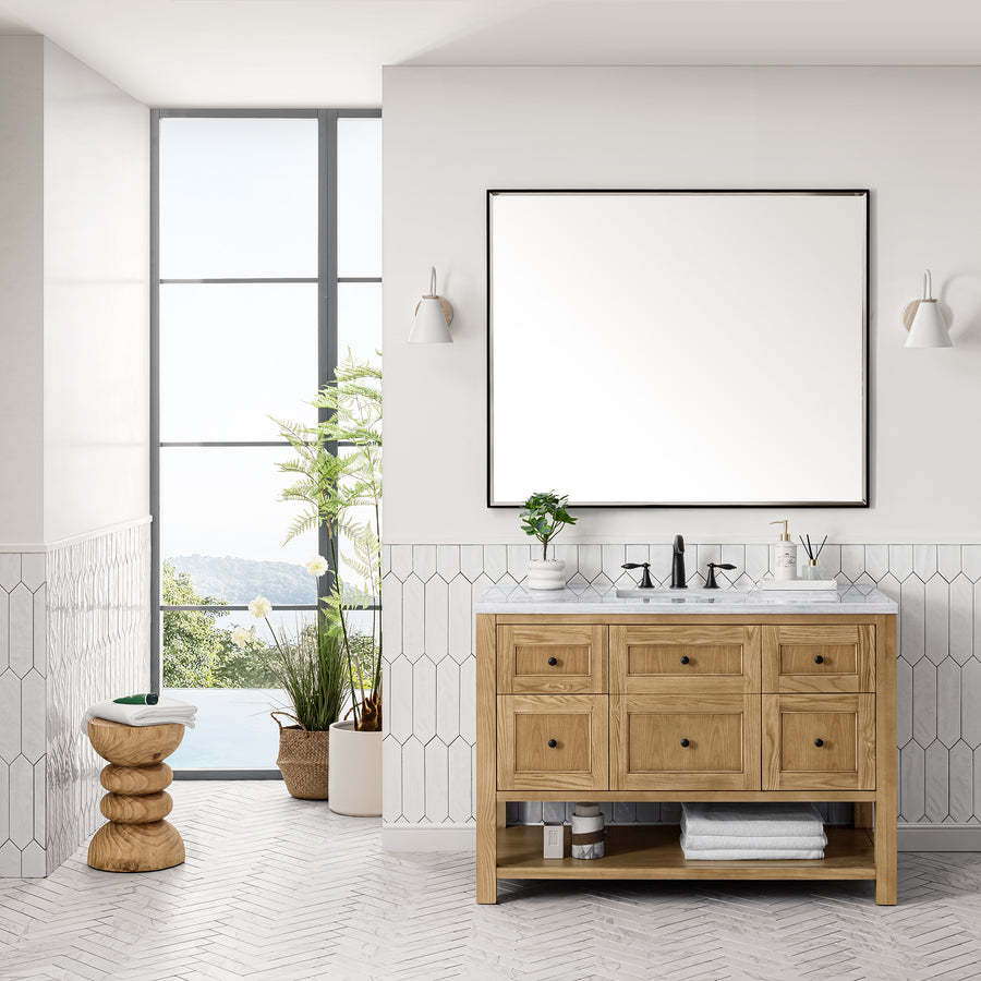 Modern bathroom featuring the Breckenridge 48" Vanity, a large mirror, white tiled walls, potted plants, and a sculptural wooden stool by a window with scenic views—showcasing luxury craftsmanship in every detail.