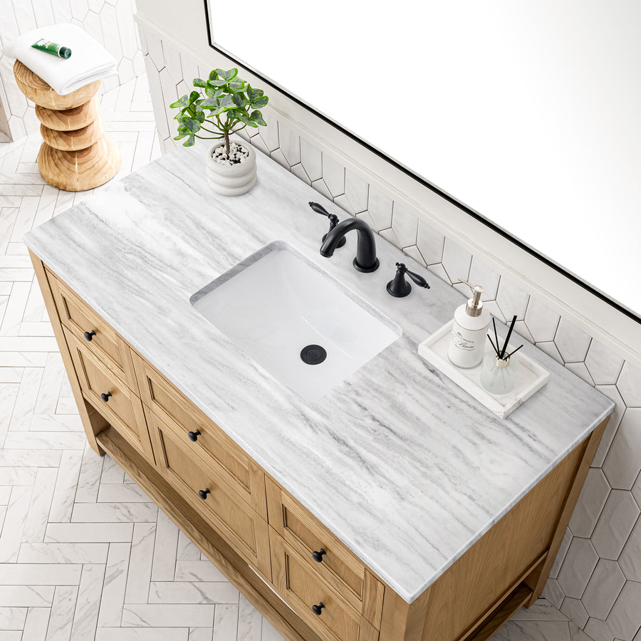 A bathroom vanity with a light wood cabinet, white marble countertop, black faucet, and a rectangular sink. On the counter are a potted plant, a soap dispenser, and a tray with a diffuser. White tile floor and wall.