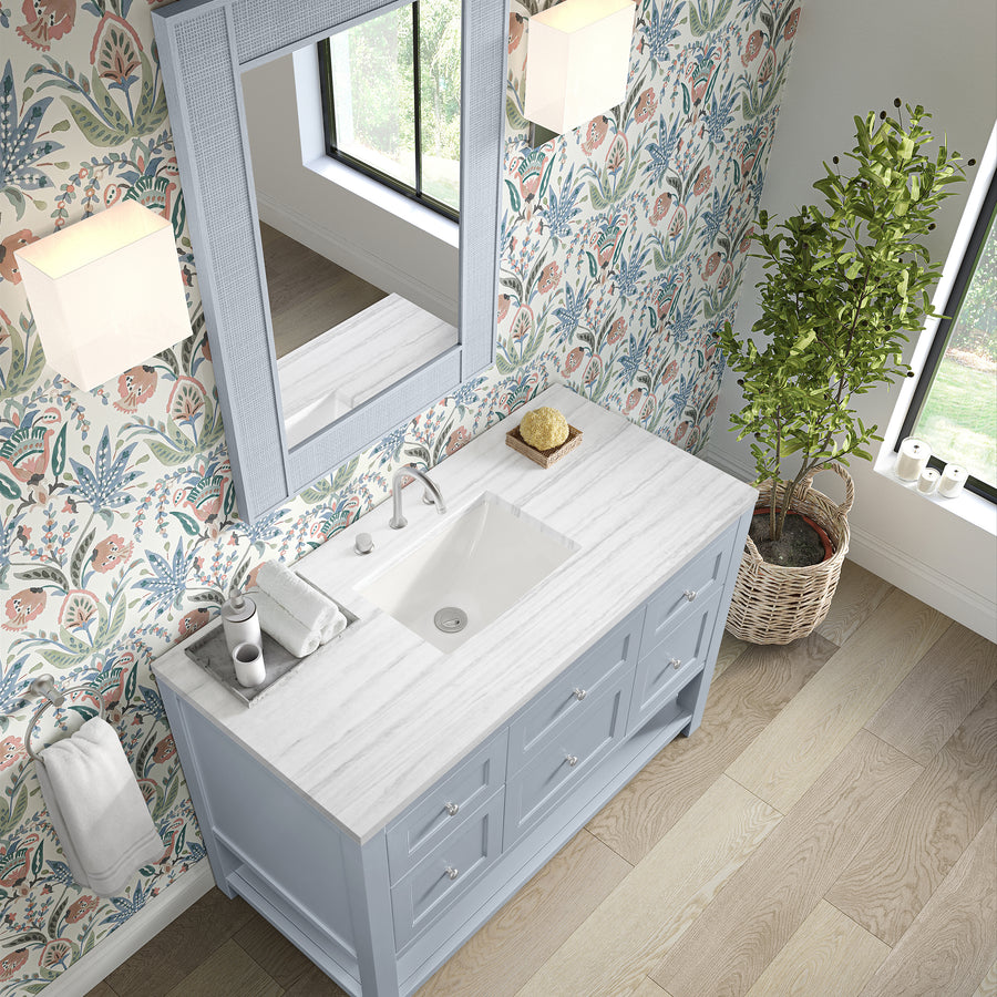 A bathroom with floral wallpaper, a large window, a potted plant, and a blue vanity with a white marble countertop, a sink, towels, soap, and a large mirror above. Light wooden flooring is visible.