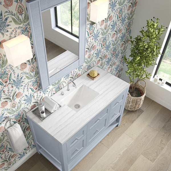 A bathroom with floral wallpaper, a large window, a potted plant, and a blue vanity with a white marble countertop, a sink, towels, soap, and a large mirror above. Light wooden flooring is visible.