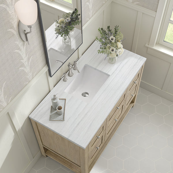 A light wood bathroom vanity with a white marble countertop, rectangular sink, silver faucet, a small tray with soap and lotion, and a glass vase with white flowers, viewed from above.