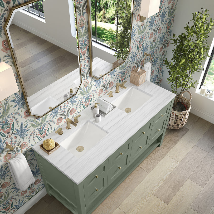 A bathroom with a green double-sink vanity, gold fixtures, floral wallpaper, two octagonal mirrors, a potted plant, and a window letting in natural light. The countertop holds soap dispensers and a tissue box.