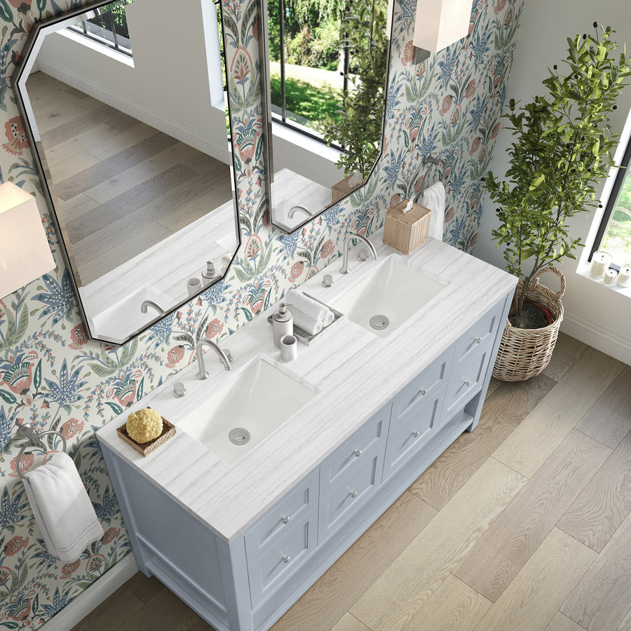Bathroom with a double sink vanity, light blue cabinets, floral wallpaper, two mirrors, a potted plant in a wicker basket, soap dispensers, and natural light from a window.
