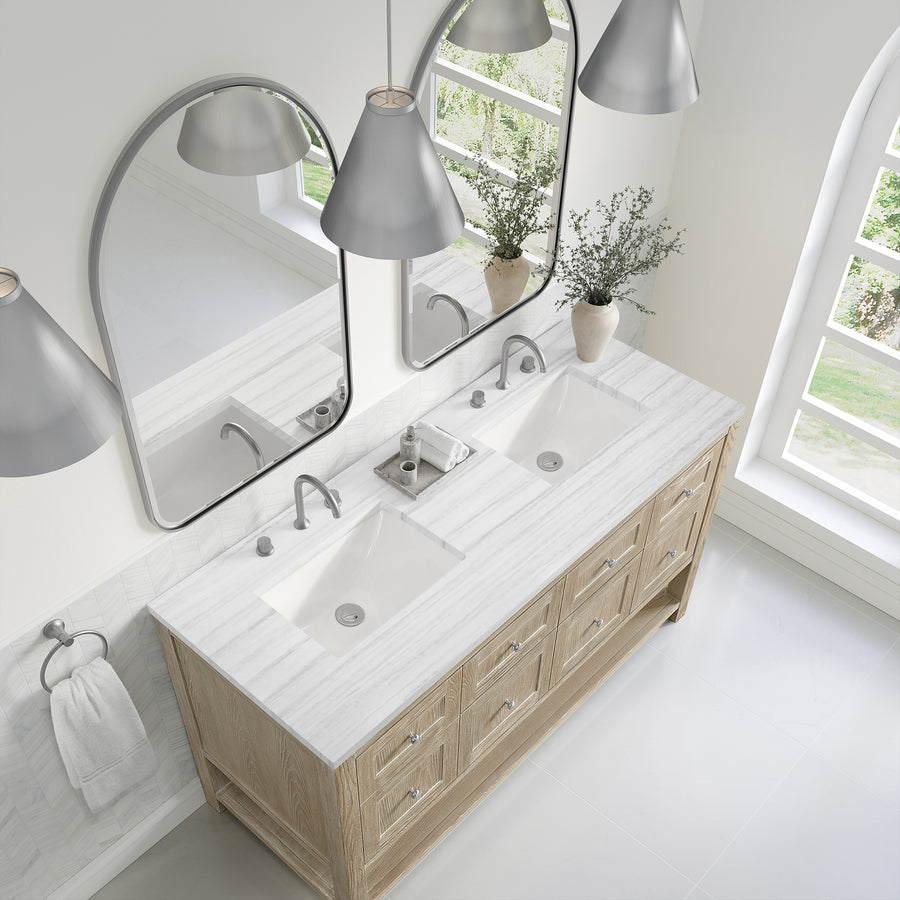 Modern bathroom with a double-sink vanity, light wood cabinets, marble countertop, two oval mirrors, silver faucets, pendant lights, a vase with greenery, and large windows letting in natural light.