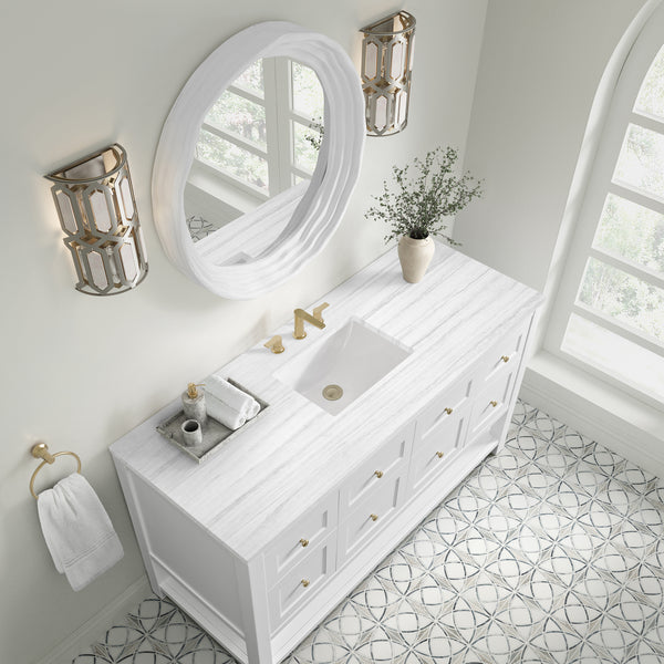 A bright bathroom with a white vanity, gold fixtures, a round mirror, modern wall sconces, a patterned tile floor, and a vase with greenery. Two windows let in natural light. A hand towel hangs on the wall.