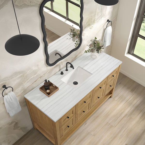 A bathroom with a wooden vanity, white marble countertop, black faucet, decorative tray, and vase. Above is an irregular-shaped mirror. White towels hang on black rings, and large windows let in natural light.