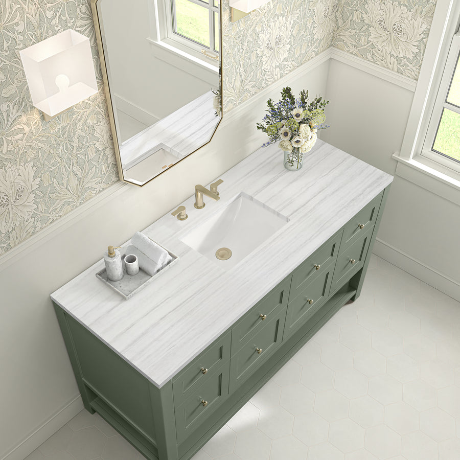 A bathroom vanity with a marble countertop, gold faucet, green cabinets, a vase of flowers, and a tray with towels and soap. A rectangular mirror and window are above, with patterned wallpaper in the background.
