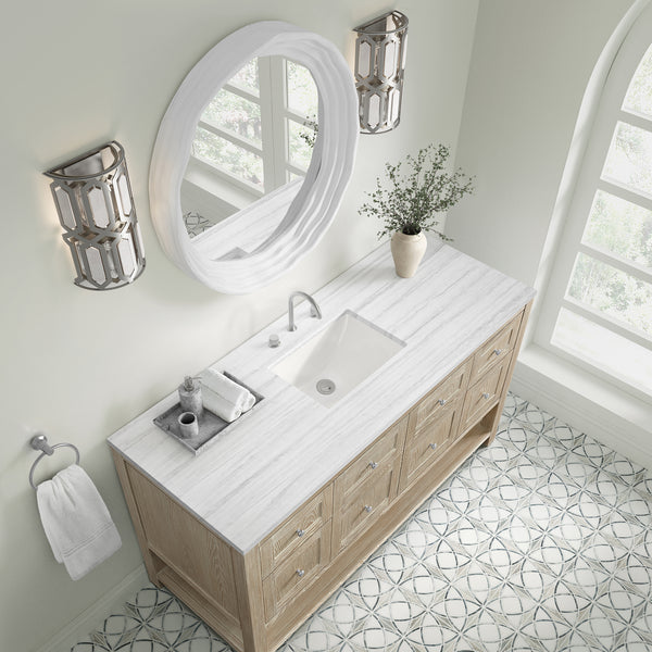 A bright, modern bathroom with a wooden vanity, white countertop, rectangular sink, oval mirror, two chrome wall sconces, potted plant, towel ring with a white towel, and patterned tile floor.