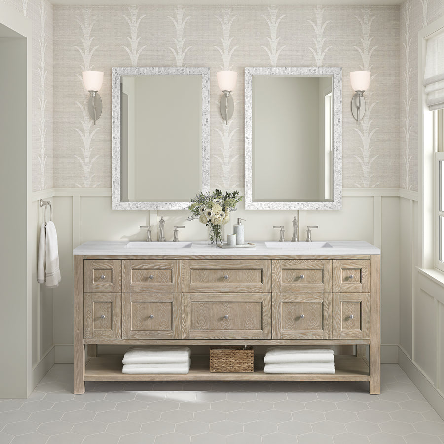 A modern bathroom features the Breckenridge 72" Double Bowl Vanity, two mirrors, wall sconces, stacked white towels, a basket below, and a vase of flowers—all set against light patterned wallpaper and neutral tones.