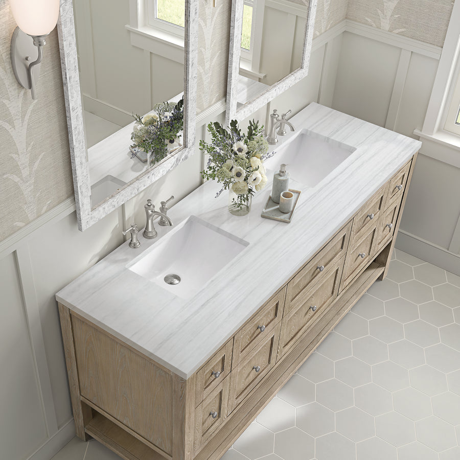 A double-sink bathroom vanity with a marble countertop, light wood cabinets, silver faucets, two large mirrors, and a vase with white flowers, viewed from above. The room has hexagonal floor tiles and soft natural light.