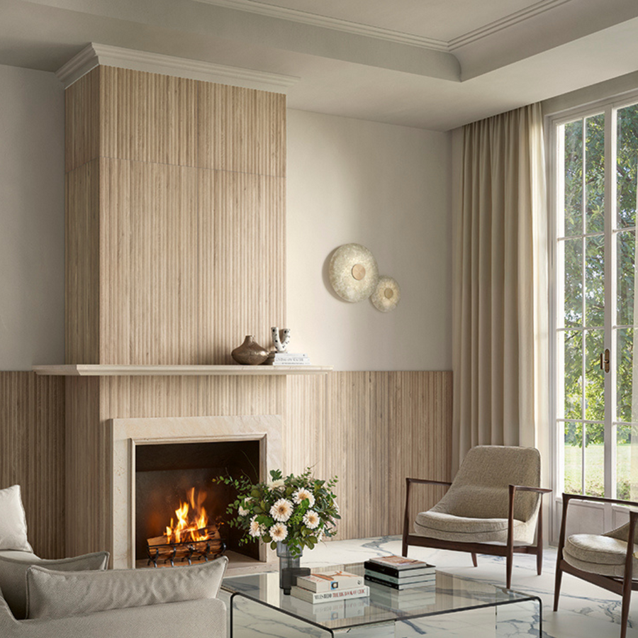 A modern living room features a wooden-paneled fireplace with Corsica Fluted 24x48 Ceramic Tile, a burning fire, armchairs, a glass coffee table with books and flowers, wall art, and large windows with cream curtains letting in sunlight.