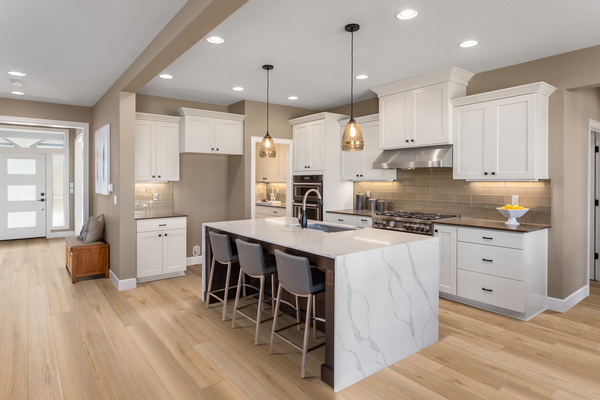 Modern kitchen with white cabinets, marble island with barstools, stainless steel appliances, pendant lights, and light wood flooring. Neutral walls and recessed lighting create a bright, spacious atmosphere.