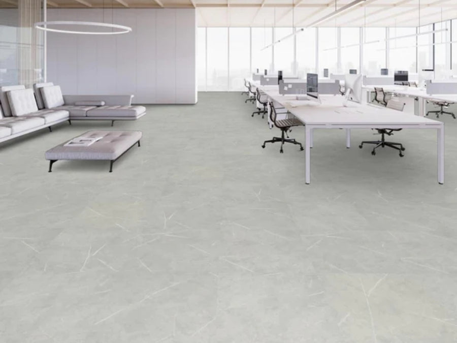 A modern open-plan office features rows of white desks, computers, and chairs to the right, a lounge with gray sofas and table to the left, large windows for natural light, and durable Commercial Vinyl flooring for a sleek finish.