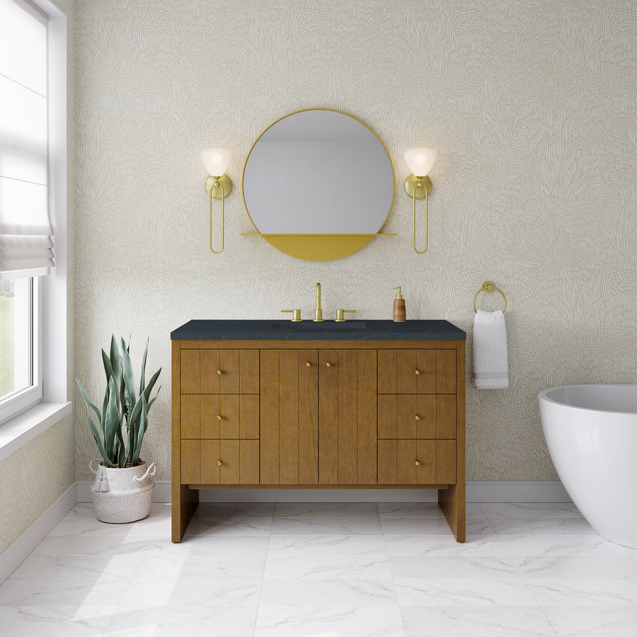 A modern bathroom showcasing the stylish Hudson 48″ Vanity in Honey Oak as its centerpiece, complemented by a round mirror, gold fixtures, wall sconces, a potted plant, a towel ring with a white towel, and a freestanding bathtub atop pristine white marble floor tiles—perfect for those seeking luxury bathroom furniture that balances style and function.