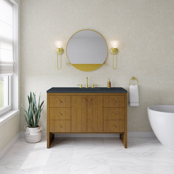 A modern bathroom showcasing the stylish Hudson 48″ Vanity in Honey Oak as its centerpiece, complemented by a round mirror, gold fixtures, wall sconces, a potted plant, a towel ring with a white towel, and a freestanding bathtub atop pristine white marble floor tiles—perfect for those seeking luxury bathroom furniture that balances style and function.