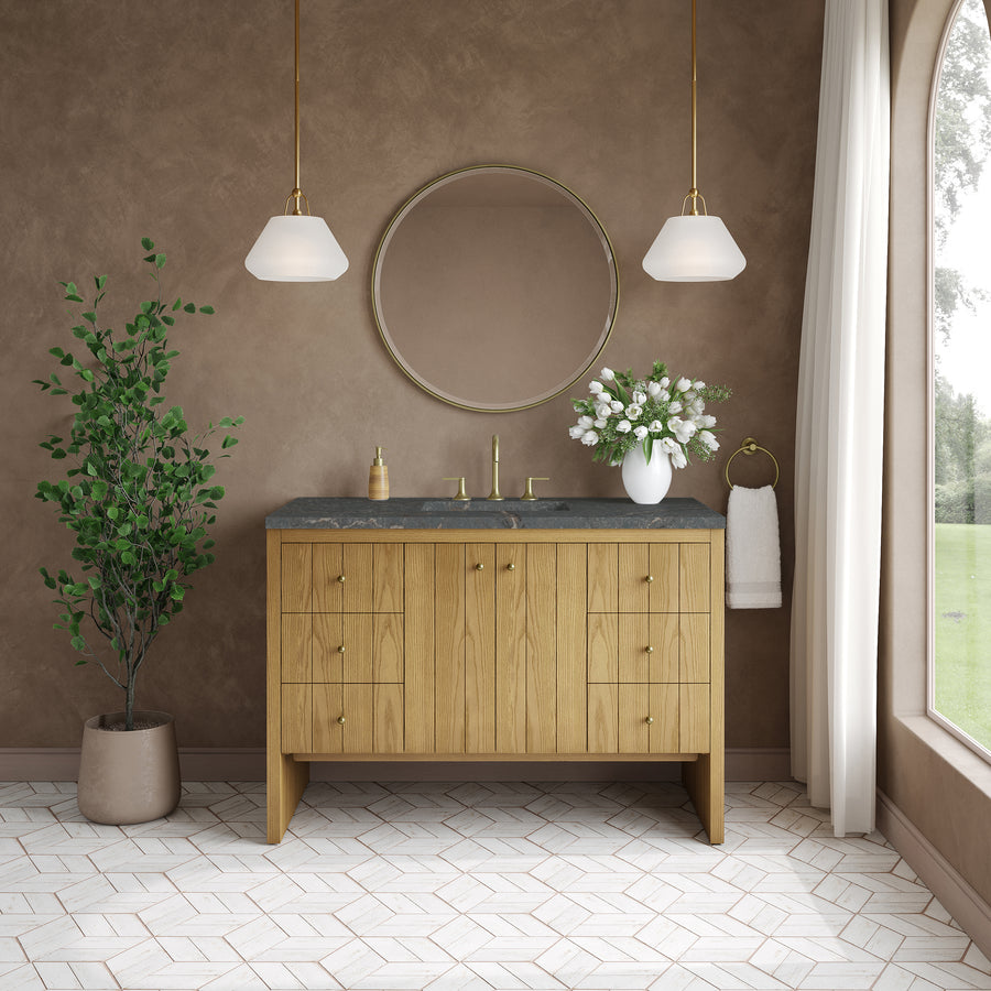 Modern bathroom featuring the striking Hudson 48″ Vanity in Light Natural Oak, complete with a sleek black countertop, round mirror, gold fixtures, two pendant lights, potted plant, vase of flowers, towel ring, and a large window adorned with white curtains that frame a picturesque view of greenery—seamlessly uniting luxurious bathroom furniture in natural oak with abundant natural light and contemporary design.