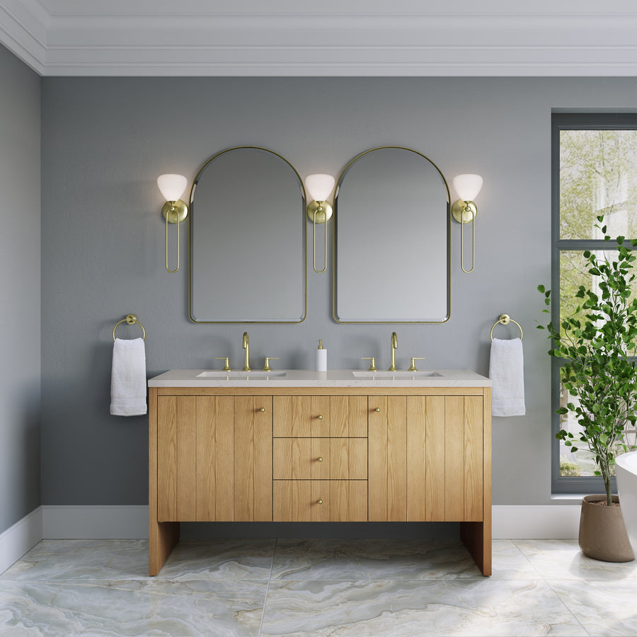 Modern bathroom featuring the Hudson 60″ Double Bowl Vanity in Light Natural Oak, double sinks, two arched mirrors with gold frames, wall sconces, towel rings, gray walls, a marble floor, and a green potted plant by the window—an inviting space that highlights luxury bathroom furniture at its finest.