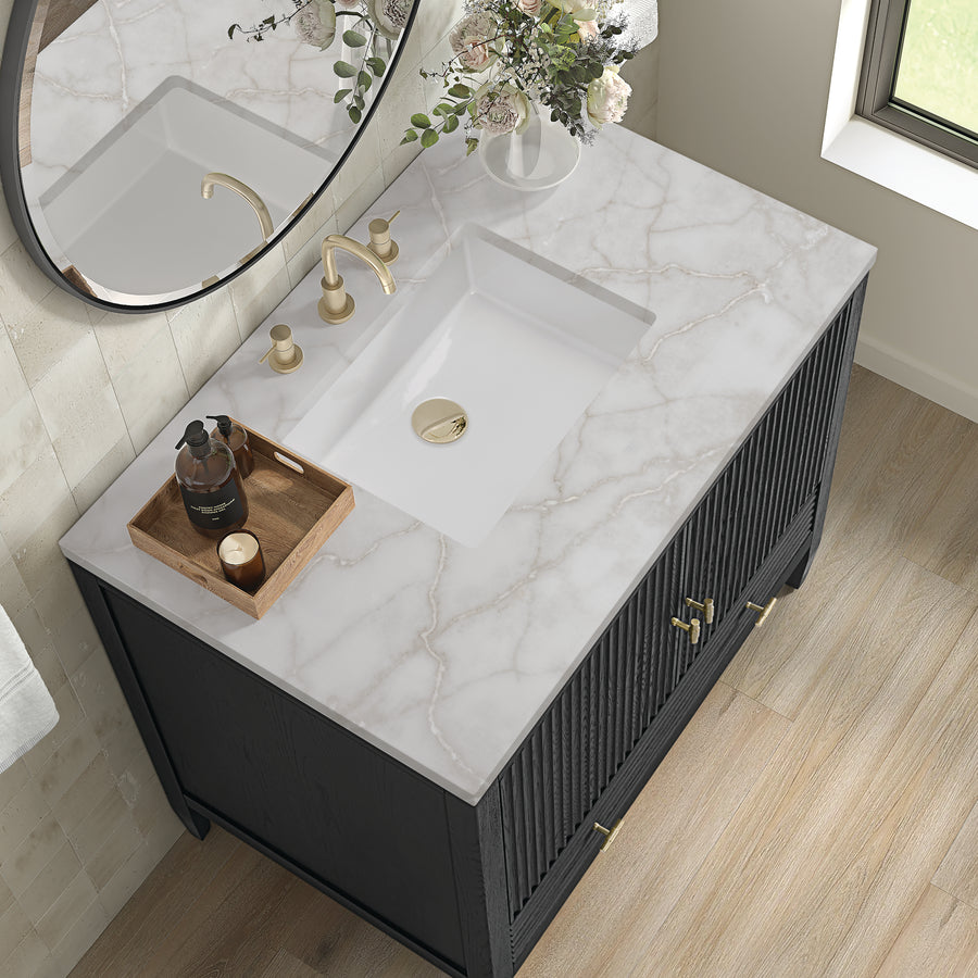 Marble bathroom vanity with a white rectangular sink, gold faucet, and handles. A round mirror, vase of flowers, and a wooden tray with toiletries sit on the countertop next to a window with natural light.