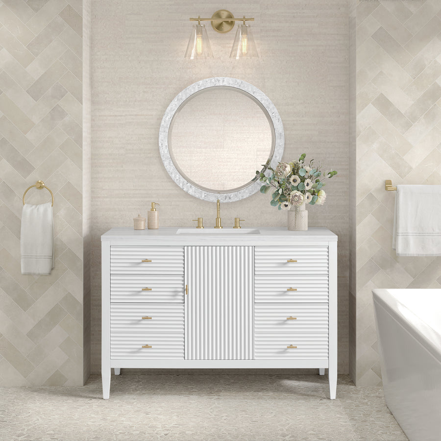 The Myrrin 48" Vanity with gold fixtures anchors the space, complemented by a round mirror and flower vase. Light beige herringbone wall tiles add warmth, while towel racks with white towels elegantly flank the 48" vanity.