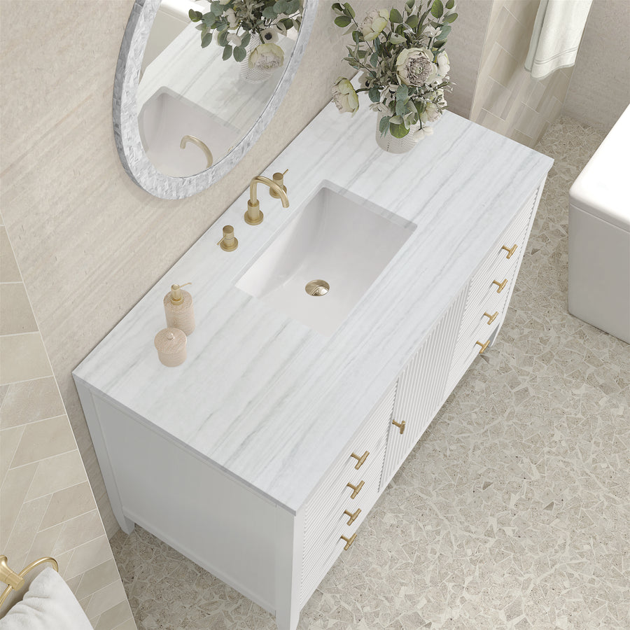 A modern bathroom with a white marble countertop, gold faucet and handles, a rectangular sink, decorative jars, a round mirror, and a vase of flowers on a light stone-tiled floor.