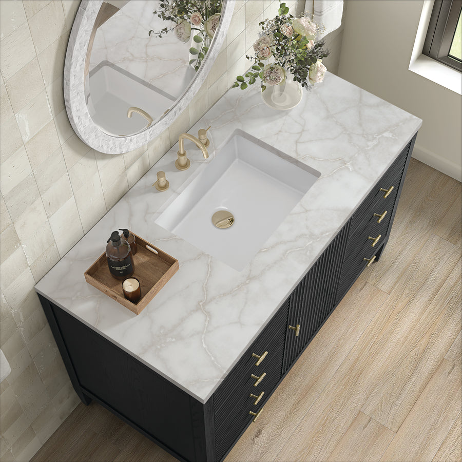 A modern bathroom vanity with a white marble countertop, rectangular sink, gold fixtures, a round mirror, a vase of flowers, and a wooden tray holding a bottle and small items, set on light wood flooring.