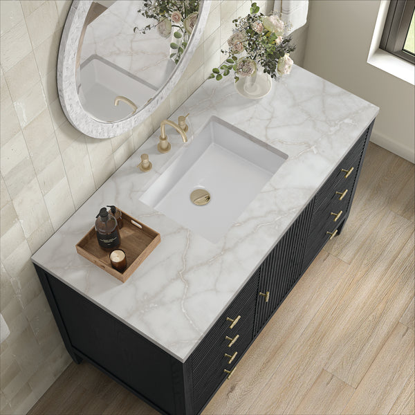 A modern bathroom vanity with a white marble countertop, rectangular sink, gold fixtures, a round mirror, a vase of flowers, and a wooden tray holding a bottle and small items, set on light wood flooring.