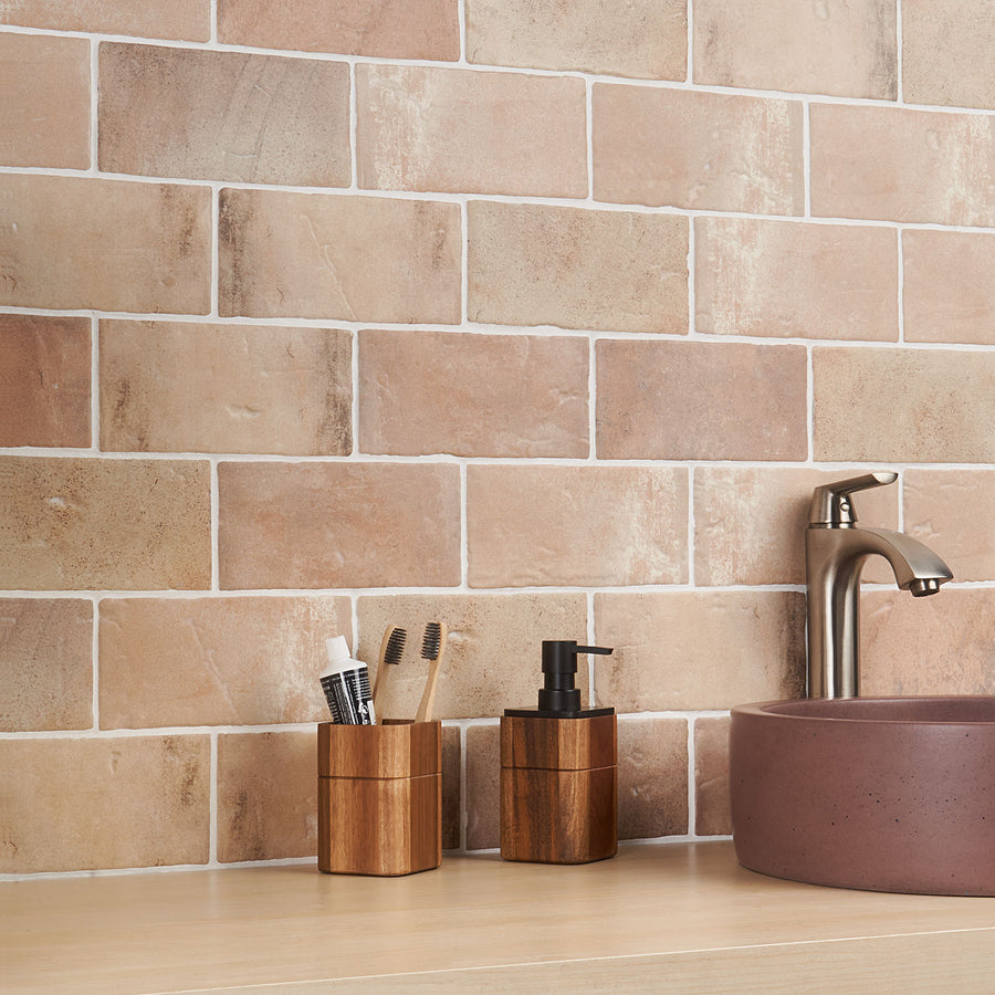 A bathroom counter with a brown sink, a silver faucet, and wooden holders containing toothbrushes, toothpaste, and a soap dispenser, set against a wall of beige rectangular tiles.
