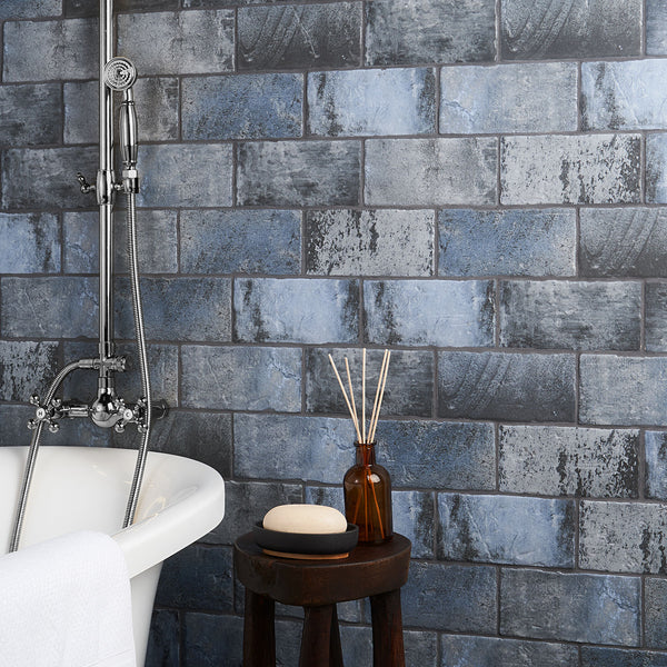 A bathroom with blue-grey brick-patterned tiles, a white bathtub with a chrome shower fixture, and a wooden stool holding a soap bar, a brown glass bottle, and reed diffusers.