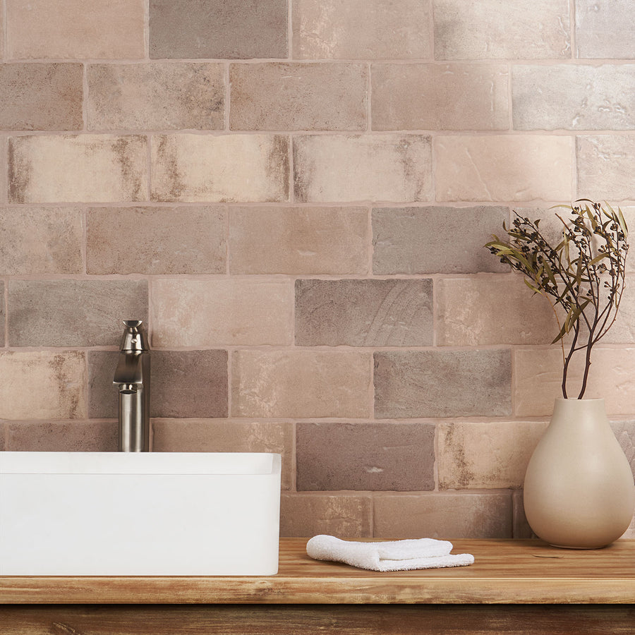 A modern bathroom countertop with a white rectangular sink, a silver faucet, a neatly folded white towel, and a beige vase holding dried branches, set against a wall of beige and brown tiles.