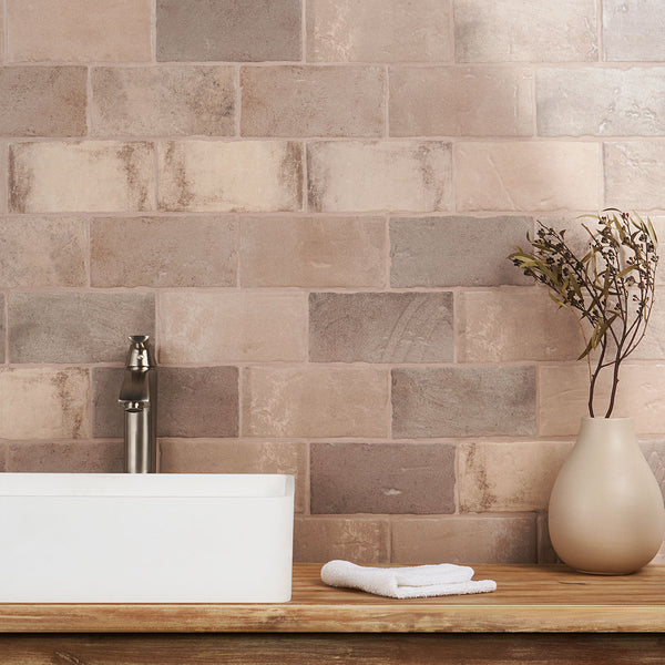 A modern bathroom countertop with a white rectangular sink, a silver faucet, a neatly folded white towel, and a beige vase holding dried branches, set against a wall of beige and brown tiles.