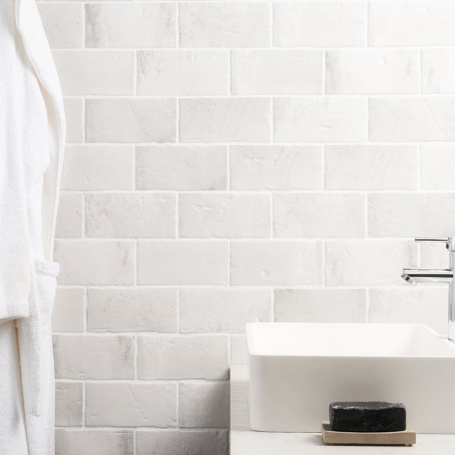 A white-tiled bathroom wall with a white sink, a chrome faucet, a black bar of soap on a dish, and a white bathrobe hanging on the left side.