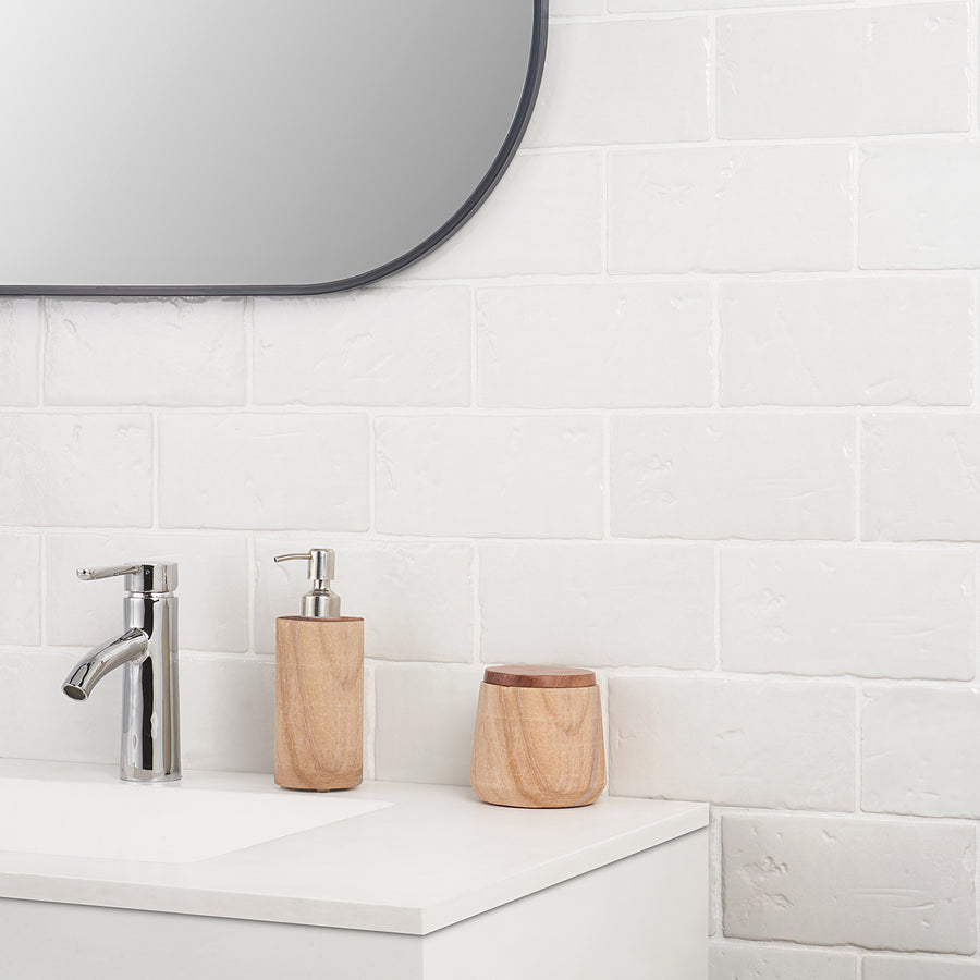 A modern bathroom countertop with a chrome faucet, a wooden soap dispenser, and a wooden container, set against a white tiled wall and beneath a rounded-edge mirror.
